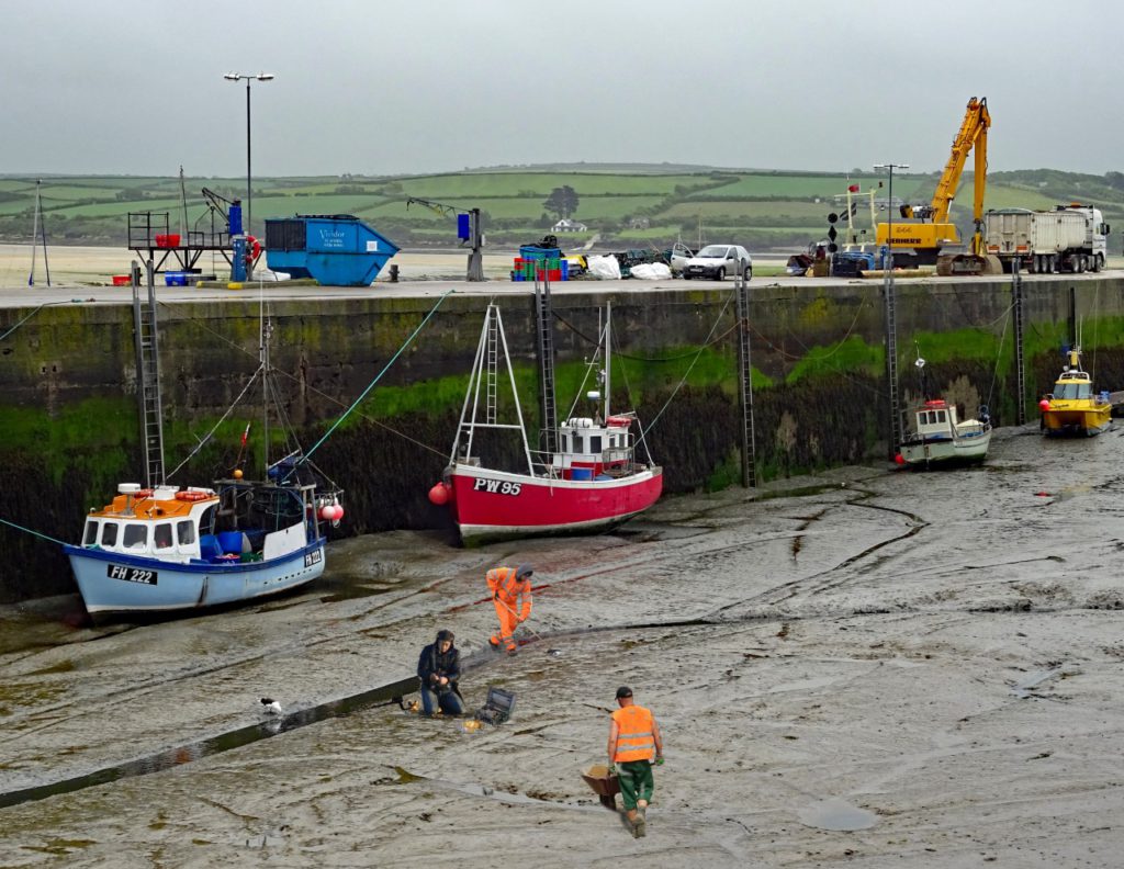 En hamn utan vatten. Längs med hamnkanten ligger det flera båtar på bottnen. På bottnet står flera personer i reflexkläder och gräver i gyttjan. Personerna ser lite konstiga ut i bilden, som att de är inklippta.